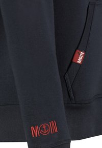 Marineblauwe sweatshirt met rode geborduurde tekst "MOIN" en ankerdesign; bevat een label van rode stof met een gestructureerd oppervlak.
