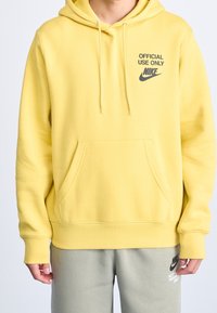 Gul luvtröja med huva och känguruficka; har svart text "ENDAST OFFICIELL ANVÄNDNING" och Nike-logotyp på övre vänstra bröstet.