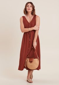 Rustfarbene, ärmellose Maxikleid mit V-Ausschnitt, Frontknöpfen, Taillenzugband und seitlichen Schlitzen. Kombiniert mit einer runden Strohtasche und Sandalen.