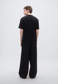 Μαύρο oversized μπλουζάκι με κοντά μανίκια και ευθεία κοπή, σε συνδυασμό με μαύρο παντελόνι φαρδιάς γραμμής, βλέποντας από πίσω πάνω σε ανοιχτό φόντο.