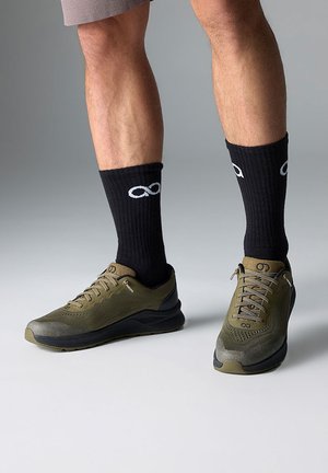 Baskets de sport vert olive avec une tige en mesh texturé et des semelles noires, associées à des chaussettes côtelées noires arborant un symbole infini blanc.