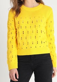 Pull jaune en tricot avec encolure ronde côtelée et motif ajouré. Manches longues avec motif texturé, associé à un jean foncé.