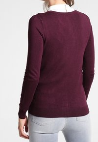 Bordowy sweter dziany z długimi rękawami i prążkowanym obszyciem. Posiada okrągły dekolt i gładką fakturę, widoczny od tyłu, bez widocznego logo.