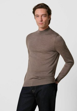 Mann trägt einen braunen Langarm-Rollkragenpullover und dunkle Jeans und steht mit einer Hand in der Tasche vor einem schlichten Hintergrund.