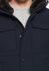 Granatowa pikowana kurtka z zamkiem błyskawicznym, dwoma kieszeniami na piersi i kołnierzem z futerka. Pod spodem teksturowany szary sweter.