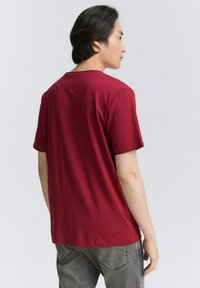 Mørkerød bomulds t-shirt med korte ærmer og rund hals, vist fra bagsiden. Har en glat tekstur og standard pasform. Grå bukser synlige.