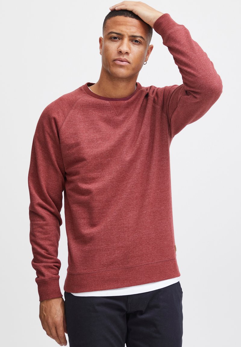 Sweatshirt rouge à manches longues, col rond et poignets côtelés. Texture douce, motif chiné subtil et une étiquette sur le côté inférieur.