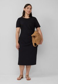 Schwarzes kurzes Kleid mit kurzen Ärmeln und taillierter Passform, das ein dezentes Drehdetail aufweist. Kombiniert mit einer gewebten beigen Tasche und flachen Sandalen in Braun.