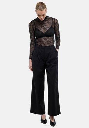 Femme aux cheveux lissés en arrière portant un haut transparent en dentelle noire à manches longues, un bralette noir, un pantalon large noir et des talons pointus noirs.