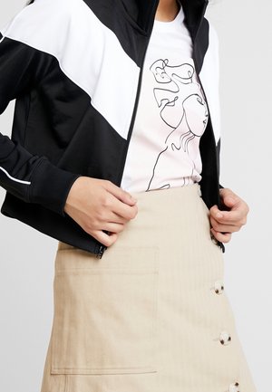 Persoon die een zwart-witte jas met rits draagt over een wit T-shirt met een zwarte abstracte gezichtslijntekening en een beige rok met knopen en een zak.
