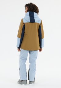 Skidjacka i brunt, blått och marinblått, med huva och kontrasterande ärmpaneler. Kombinerad med ljusblå skidbyxor och grå stövlar.