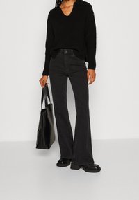 Donna che indossa un maglione nero a coste, jeans neri a vita alta a zampa d'elefante, scarpe col tacco nere e tiene una grande borsa a tracolla in pelle nera.