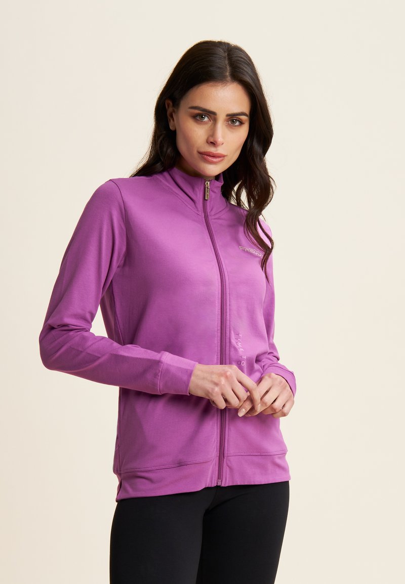 Giacca viola con zip, colletto alto, maniche lunghe e texture liscia. Presenta una zip decorativa e un branding minimale sul davanti.