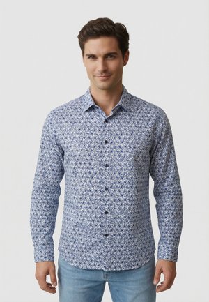 Hombre vistiendo una camisa de manga larga abotonada con patrones florales azules y jeans azul claro, posando frente a un fondo blanco liso.