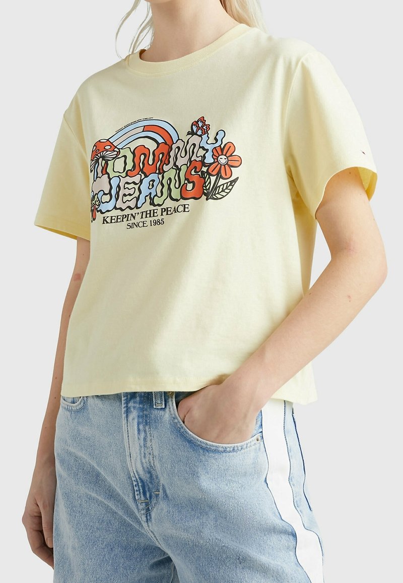 T-shirt court jaune avec un graphisme coloré de fleurs, un arc-en-ciel, et le texte "Tommy Jeans, Keepin' The Peace Since 1985." En matière coton.