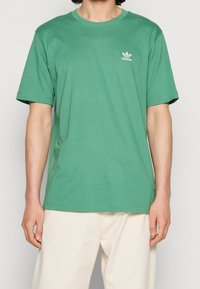 Grön bomullströja med korta ärmar, rund halsringning och en liten vit Adidas-logotyp på vänster bröst. Enkelt, avslappnat snitt.