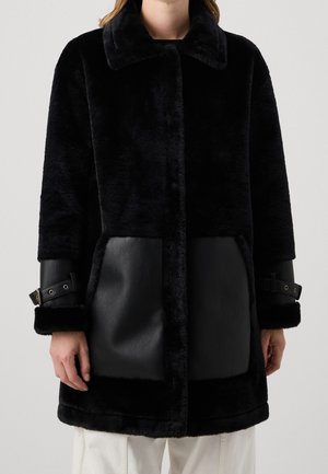 Zwarte faux fur jas met brede kraag, leren zakken als patches en gespdetails op de manchetten, gedragen over witte broeken.