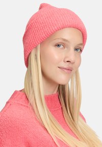 Blonde vrouw met een roze gebreide beanie en een bijpassende roze trui, lichtjes glimlachend, tegen een lichtgrijze achtergrond.