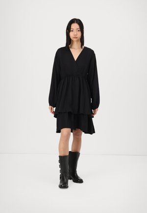 Black dress with a tiered design, long sleeves, and a deep V-neck. Worn with knee-high black boots featuring buckle accents.