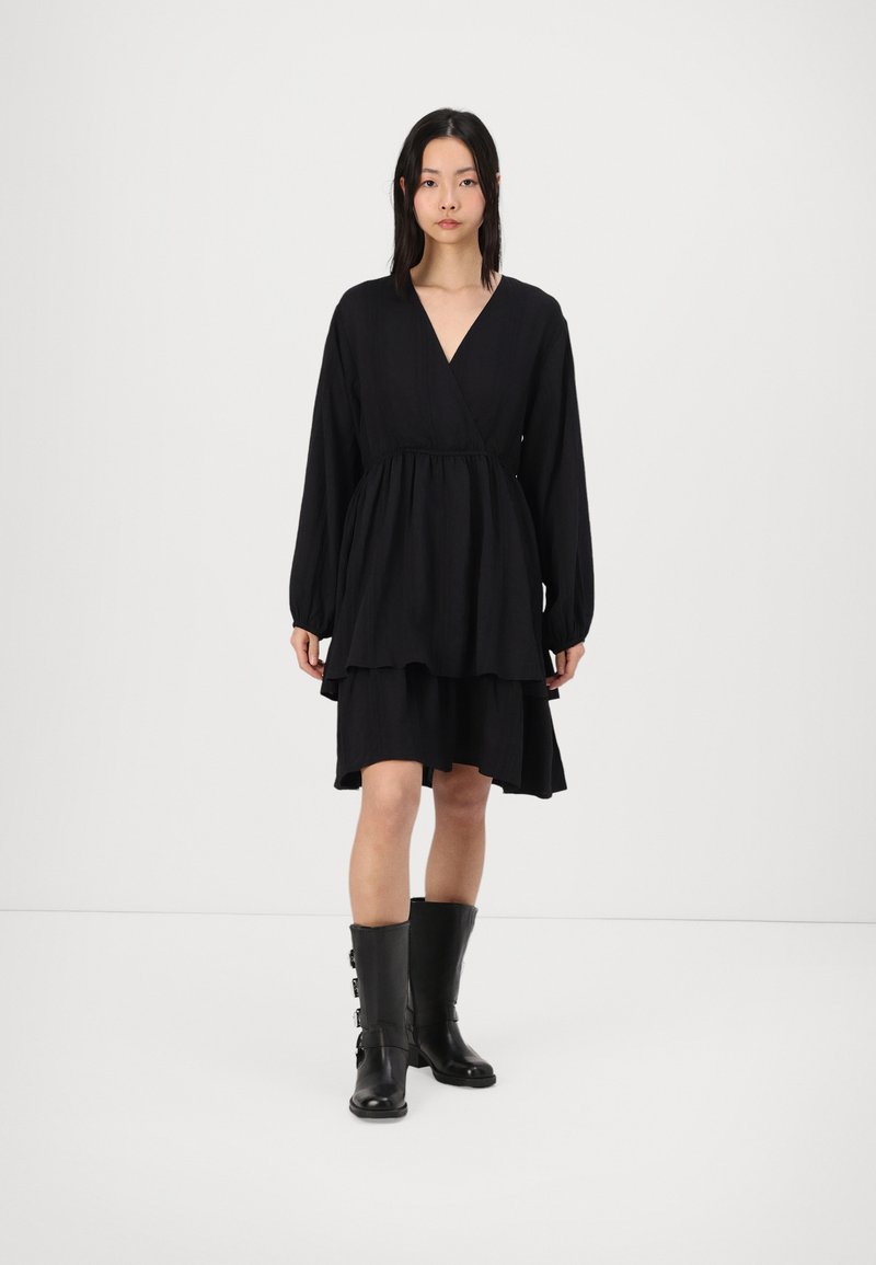 Black dress with a tiered design, long sleeves, and a deep V-neck. Worn with knee-high black boots featuring buckle accents.
