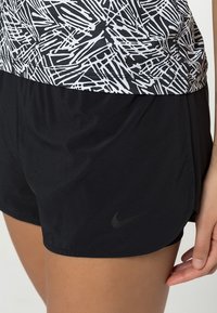 Primer plano de una persona que lleva puesta una camiseta de patrón abstracto en blanco y negro y pantalones cortos deportivos negros de Nike, con la mano descansando al lado de los pantalones.