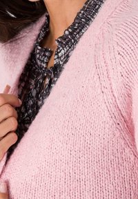 Ljusrosa, kraftig stickad cardigan med texturerad yta, lagd över en blus med svart och vitt mönster som har en volanghalsringning.