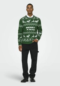 Maglione di Natale verde a maglia con renne bianche, fiocchi di neve e testo "BUON NATALE", abbinato a pantaloni neri e scarpe nere.