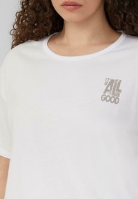 T-shirt en coton blanc avec un col rond et des manches courtes. L'avant présente un graphique texturé gris avec le texte "C'EST TOUT BON."