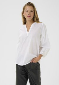 Weißer Pullover-Bluse mit V-Ausschnitt und langen, gerafften Ärmeln. Der strukturierte Stoff weist dezente vertikale Streifen auf. Wird mit schwarzen Hosen getragen.