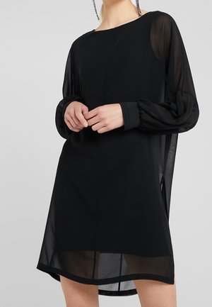 Frau trägt ein schwarzes, durchsichtiges langärmliges Kleid mit gepufften Ärmeln, steht mit vor dem Körper verschränkten Händen vor einem einfachen Hintergrund.