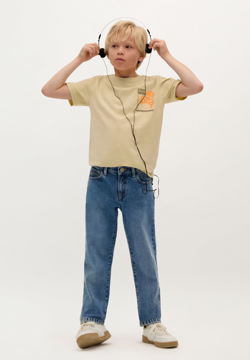 Korte mouw lichtbeige t-shirt met een schedelafbeelding, gecombineerd met blauwe denim jeans en witte sneakers, met zwarte hoofdtelefoon op.