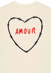 Kreemit värvi T-särk, millel on õmmeldud südame kontuur ja sõna "AMOUR" julgete punaste tähtedega südame kujundis.