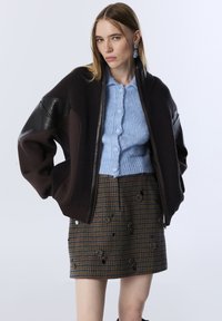 Jonge vrouw in een oversized donkerbruine jas, een lichtblauwe knooptrui, een gemusterde minirok met versieringen en oorbellen die hangen.