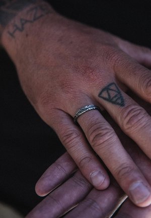 Silver ring with a textured, twisted design on a hand with a visible tattoo; skin tone varies, hands are positioned together.