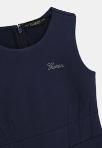 Marineblaues ärmelloses Kleid mit rundem Ausschnitt, verziert mit einem silbernen Strass-"Guess"-Logo und dezenten Falten im Rock.