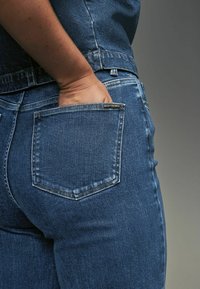 Mörkblå denim jeans med rak passform, som visar en bakficka och sömnadsdetaljer. Tyget ser texturerat ut med en aning blekning.