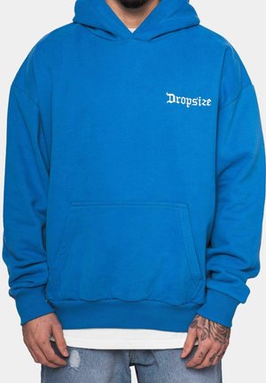 Man wearing bright blue hoodie with white "Dropsize" text on chest, paired with light blue distressed jeans and hand tattoos visible.