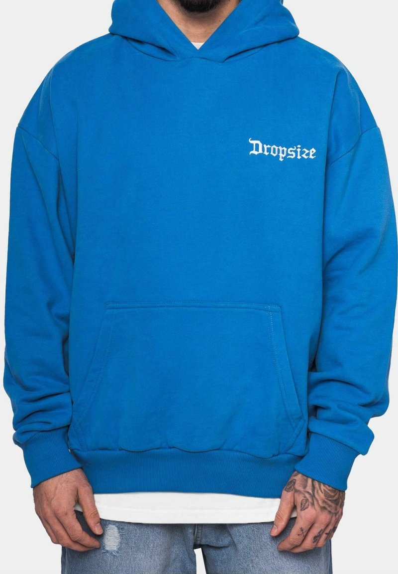Homme portant un sweat à capuche bleu vif avec un texte blanc "Dropsize" sur la poitrine, associé à un jean délavé bleu clair et des tatouages visibles sur les mains.