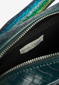 Interno della borsa intrecciata color teal con fodera nera, cerniera argentata e un'etichetta che recita "LOVE & ROSES." Presenta una tracolla multicolore e scintillante.