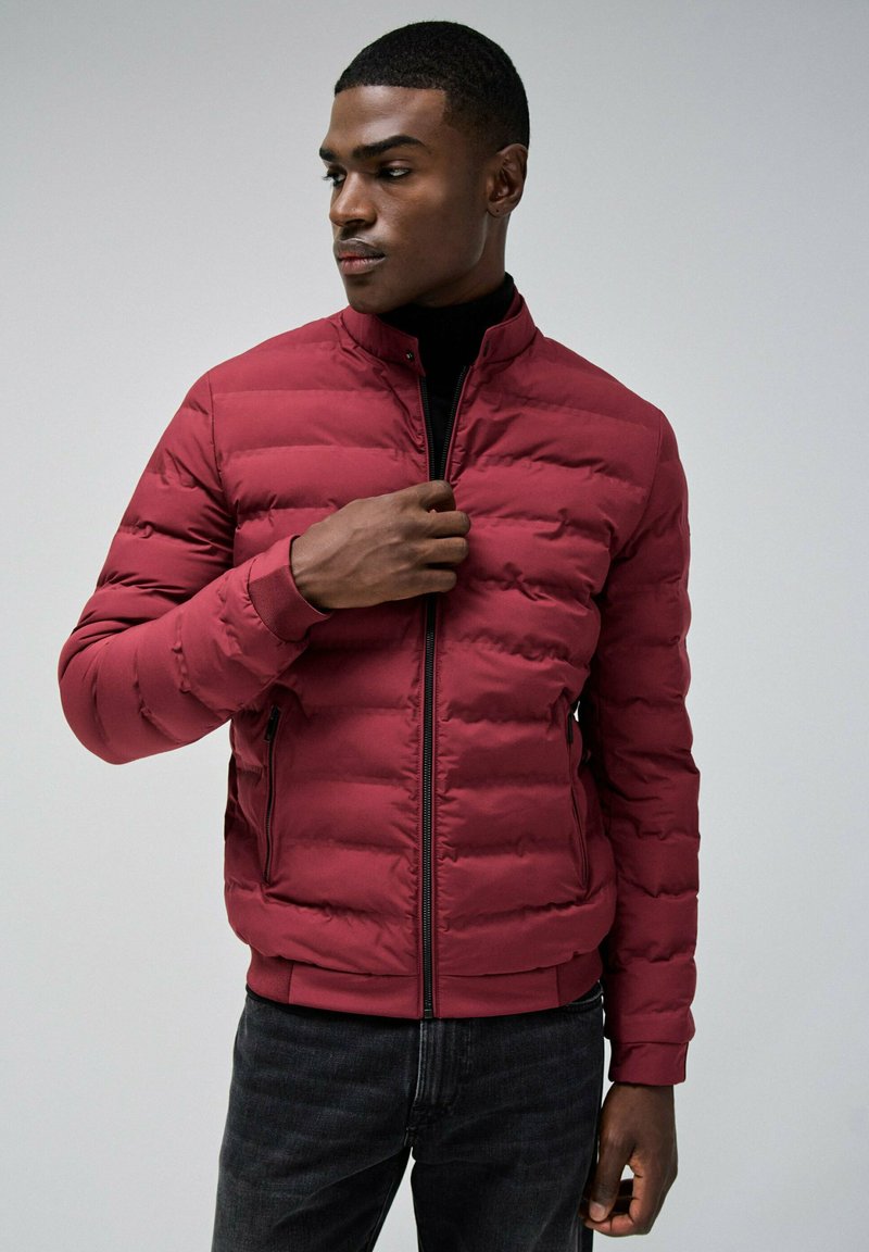 Chaqueta acolchada burdeos con diseño acolchado, cuello alto, cremallera frontal y bolsillos laterales. La tela parece suave con un ligero brillo.