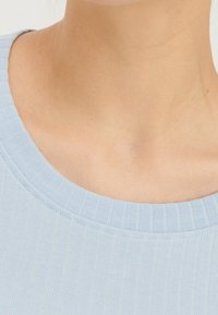 Närbild av en persons nacke och övre bröst med en ljusblå ribbad stickad topp med rund halsringning.