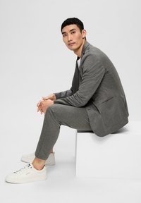 Graue, strukturierte Jacke mit passenden Slim-Fit-Hosen, kombiniert mit weißen Sneakern. Besitzt eine einzige Tasche an der Jacke und klare Linien.