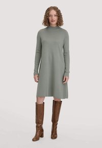 Knie-langes, langärmliges Kleid aus hellgrünem Strickstoff mit hohem Kragen. Kombiniert mit braunen, ledernen kniehohen Stiefeln.