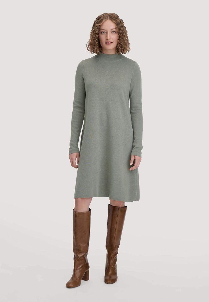 Knie-langes, langärmliges Kleid aus hellgrünem Strickstoff mit hohem Kragen. Kombiniert mit braunen, ledernen kniehohen Stiefeln.
