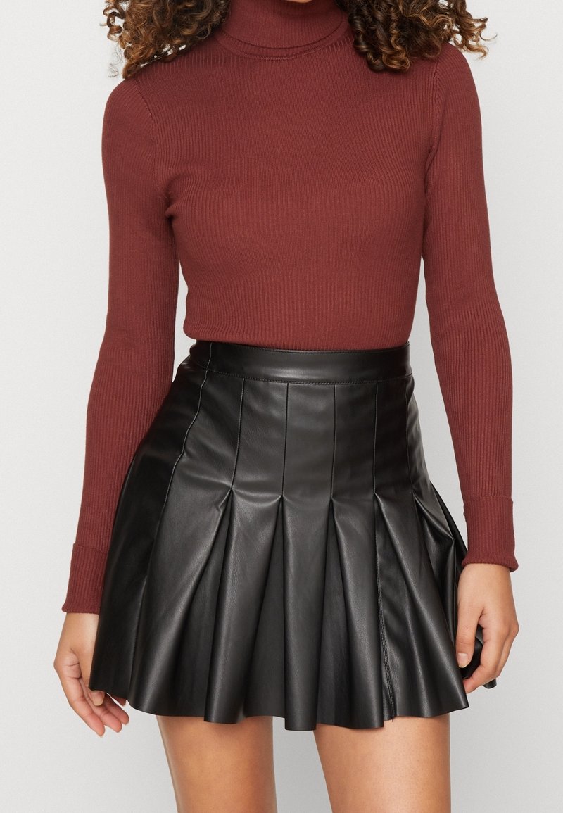 Femme portant un col roulé côtelé bordeaux et une mini-jupe plissée en cuir noire, debout devant un fond uni.