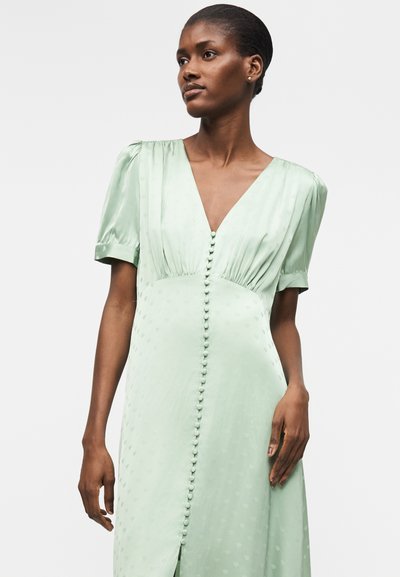 Γυναίκα που φοράει ανοιχτοπράσινο, κοντομάνικο φόρεμα με κουμπιά καλυμμένα με ύφασμα μπροστά και βαθύ V-λαιμό, στέκεται μπροστά σε απλό φόντο.
