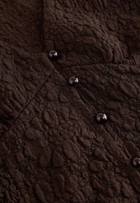 Dark brown textured fabric with a raised, patterned design, featuring small glossy round buttons along a seam.