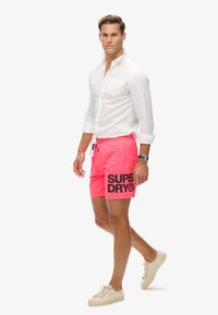 Roze zwemshorts met het "SUPER DRY" logo in het zwart op het linkerpijpje; gecombineerd met een wit overhemd met knopen en beige sneakers.