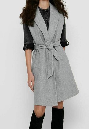 Femme portant un manteau sans manches gris ceinturé, par-dessus une chemise noire aux manches retroussées et des bottes noires jusqu'aux genoux, debout devant un fond uni.