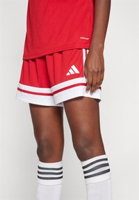 Červené atletické šortky s bielymi horizontálnymi pruhmi a logom Adidas. V kombinácii s bielymi ponožkami po kolená so čiernymi pruhmi.